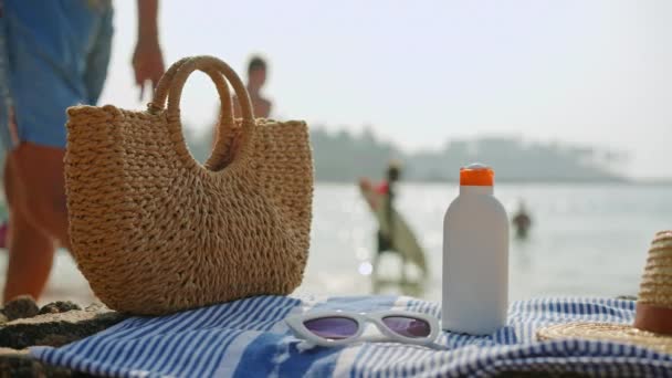 家族は海を楽しみ バックグラウンドで遊びます サンクリームボトル 編まれたバッグ ストライプタオルのサングラス付きのビーチシーン サマーレジャー 晴れた海岸でのスキンケアルーチン — ストック動画