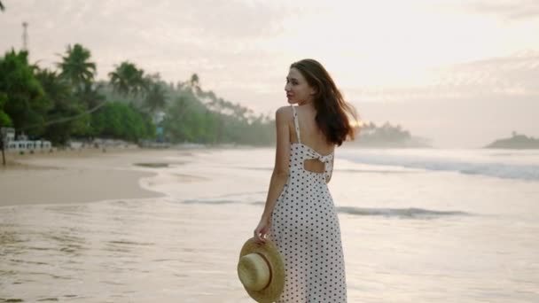 ドレスを着た美しい笑顔の女の子が回り 海岸を歩き 日の出にサーフィンをします 若い白人女性は 空の海岸に沿って 熱帯植物に沿って行きます 休暇でリラックスする幸せな女性 — ストック動画
