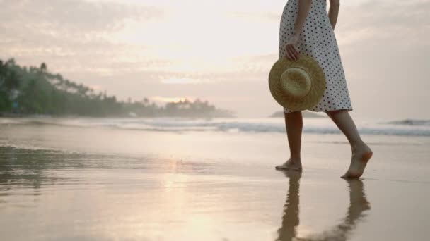 日の出に海水の端でリラックスして歩く女性の足は 水に反射します 女性の足は 夏の朝に海岸沿いに裸足を歩いています ドレスの女の子 ストロー帽子を保持 休暇を楽しむ — ストック動画