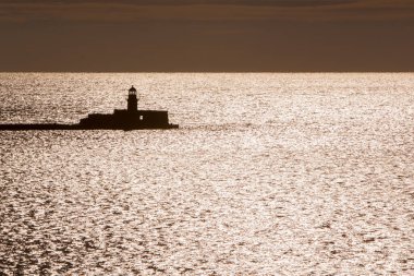 Sakin suların üzerinde sakin günbatımı, sahil fenerinin silueti. Huzurlu deniz sahnesi güvenlik ve rehberlik gerektirir..