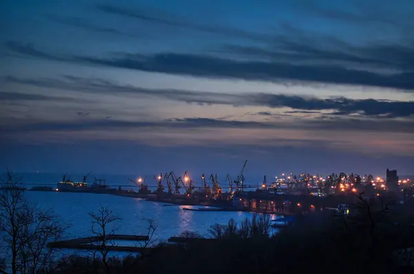 Maritime industrial hub transitions to night, highlighting harbor activity and urban backdrop with emerging city lights. Twilight descends on busy port with cranes silhouetted against dusk sky.