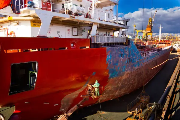Maritime vessel maintenance, industrial painting job at shipyard. Worker paints cargo ship hull red in dry dock under blue sky. Large tanker refurbishment, steel boat repair work, sunny day.