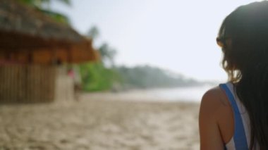 Mutlu kız bungalovlu kumsalda hindistan cevizi kokteyli içiyor. Genç, güzel bir kadın dinleniyor, tropik sahilde güneşleniyor. Güneş gözlüklü gülümseyen blogcu güneşli yaz gününde deniz kenarında güneşleniyor.