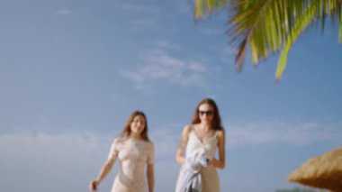 Mavi gökyüzü, güneşli yaz gününde palmiye ağacı. İki genç kadın konuşuyor, tropik plajda plaj battaniyesini sallıyorlar. Neşeli beyaz elbiseli kızlar rahatlar, sahilde dinlenirler. En iyi arkadaşlar sahilde dinlenir..
