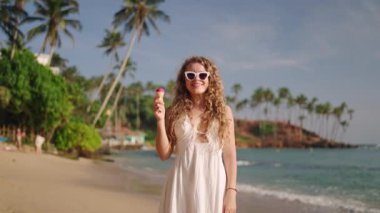 Sıradan bir yaz elbisesi, güneş gözlükleri, dalgalar, arka planda palmiye ağaçları. Gülümseyen kadın sahilde gezer, dondurmalı tropikal manzarayı sever. Yürüyüş, tatlı, güneşli deniz manzarası. Yavaşla