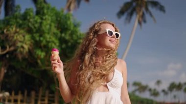 Kıvırcık saçlı kadın tropik plajlarda geziniyor, gün ışığında dondurmanın tadını çıkarıyor. Dondurmalı tatlı, sıcak iklimli atmosfer, tasasız. Keyif yürüyüşü, şekerleme, esintili sahil manzarası..