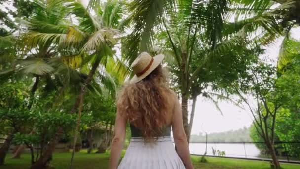 卷曲的头发游客欣赏热带的树叶 宁静的湖景 戴草帽的女人探索茂密的植物园 游人享受宁静的大自然 绿树成荫的环境 — 图库视频影像