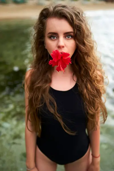 Natural beauty, wellness, summer fashion concept. Attractive woman in swimsuit holds red hibiscus in mouth by water. Feminine allure with tropical flora. Lady enjoys beach lifestyle, relaxation.