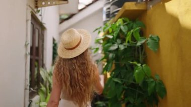 Genç kıvırcık kadın yürüyor, Bali 'de yeşil bitkilerle dar cadde boyunca dönüyor. Gezgin blogcu gider, tropikal adadaki egzotik bitkilere dokunur. Güzel gülümseyen bayan tatilde yürümekten hoşlanır..