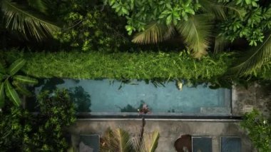 Kokteyl içer, tropik cennette yalnızlığın tadını çıkarır. İHA 'lı bikinili kadın. Etrafı yeşilliklerle çevrili, berrak bir havuzda uzanıyor. Güneş ışığı palmiyelerin arasından süzülür, suyu yansıtır..