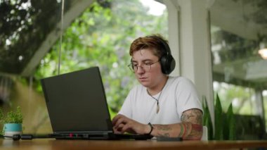 Ekrana odaklanmış, kulaklık takıyor, dizüstü bilgisayardaki klipleri düzenliyor. Dövmeli transseksüel editör çevre dostu çalışma alanında video projesinde çalışıyor. Hoş geldin, çeşitli uzak çalışma ortamı.