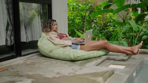 自由职业者喜欢远程工作 周末在绿色植物中休闲 在热带花园的笔记本电脑上工作的是松驰的桦树女人 在豆袋边的游泳池上悠闲地躺着 慢动作 — 图库视频影像