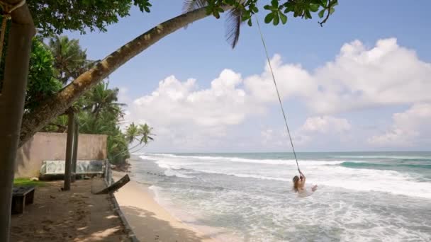 在阳光衣服 棕榈树背景 海风的欢乐时刻 女人在热带海滩波浪上方的绳索上摆动 在清澈的水面上荡秋千 冒险旅行 在异国情调的海岸线上进行休闲活动 — 图库视频影像
