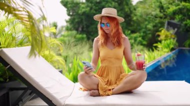 Günlük iş kavramı, boş zamanı tropikal ortamdaki işlerle harmanlamak. Sarı elbiseli kızıl saçlı kadın mavi havuz kenarında dinleniyor, karpuz kokteyli ile yaz gününün tadını çıkarıyor, akıllı telefondan mesajları kontrol ediyor..