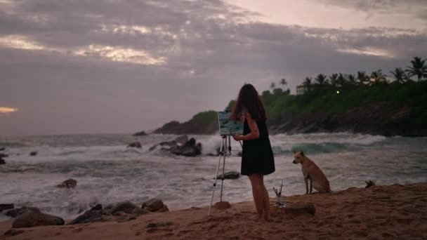 旁边的狗友 海浪拍打着岩石 黄昏时分 女画家在帆布上的汹涌大海中描绘海景 富有创造力的女性在流淌 自然背景下 在日落海滩上激励着艺术创作 慢动作 — 图库视频影像