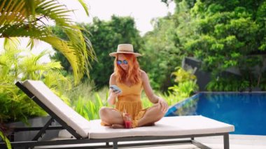 Yaz tatili, serbest çalışma, tropikal bir his. Sarı elbiseli kızıl saçlı kadın havuz başında gevşiyor, kokteylinden yudumluyor, akıllı telefona bakıyor. Teknoloji dengesi, güneşli otel ortamında ferahlama. Yavaşla