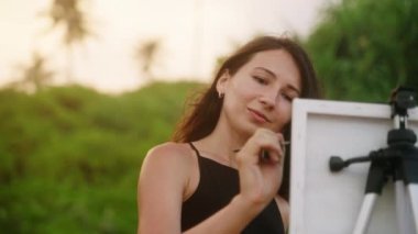 Fırçayla yaratıcı sanatçı, kumsalda sehpa. Gün batımında kadın tuval üzerinde resim yapıyor. Kişisel gelişim ve rahatlama için sanat hobisi. Palmiye ağaçları, sakin doğada sıcak bir ışık. Yavaş çekim.