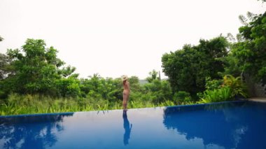 Yan manzara, tropikal manzaranın tadını çıkarmak, açık gökyüzünün altında tempoyla yürümek. Mayo giymiş bir kadın lüks villada sonsuz havuz kenarında yürür. Seyahat lüksü, huzurlu tatil inzivası. Yavaş çekim.