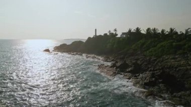 Yol gösterici gemilere rehberlik ediyor, açık gökyüzünün altında, yemyeşil yaprakların arasında ikonik deniz simgesi olarak duruyor. Hava görüntüleri deniz fenerini kayalıkların tepesinde yakalıyor, dalgalar aşağıdaki kayalık kıyılara çarpıyor..