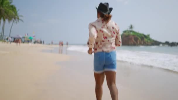 目の接触のLgbtq個人を祝い デニムショーツは熱帯の海岸を楽しんで 包摂的な旅行 夏のバイブを促進します カウボーイの服装の活気に満ちた男は岸に沿って走り ビーチの休日のために視聴者を招待します — ストック動画