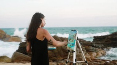 Dalgalar çarpar, gündelik elbiseli kadın resim yapmaktan hoşlanır, okyanus güzelliğini yakalar. Dişi ressam kayalık sahilde deniz manzarası çiziyor. Sanat yaratımı, manzara, dingin doğada ilham. Yavaşla