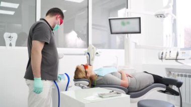 Modern diş kliniğinde profesyonel UV diş beyazlatma prosedürü. Erkek dişçi lazer diş beyazlatma sırasında kadın hastayı kontrol ediyor. Koruyucu kırmızı gözlüklü sarışın kadın. Kozmetik dişçiliği.