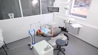 Modern diş kliniğinde profesyonel ultraviyole diş beyazlatma prosedürü. Lazer diş beyazlatma sırasında kadın hasta. Modern diş muayenehanesinde koruyucu kırmızı gözlüklü sarışın kadın. Kozmetik dişçiliği.