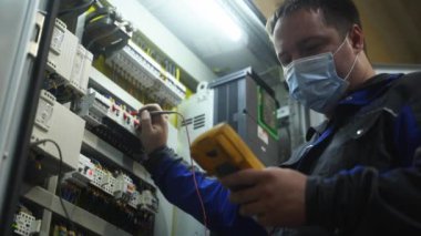2021-09-23 - Mariupol, Ukrane. Magma fabrikası. Maskeli işçi ekipmanları endüstriyel ortamda ayarlar, sistem bütünlüğünü sağlar. Odaklanmış elektrikçi otomasyon dolabını çoklu metre ile test etti.