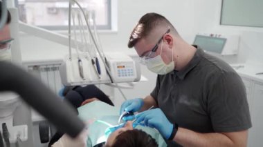 Modern diş hekiminde çalışan dişçi ve asistan. Doktor dişlerindeki taşları ve sert plağı çıkarıyor. Asistanın ağzında tükürük püskürtücü var. Stomatolog hastayı ameliyata hazırlayın..