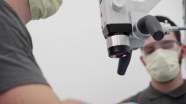Modern diş kliniğinde ameliyat sırasında diş mikroskobu kullanan erkek dişçi ve asistan hastaların yakın çekimleri. Endodontist, kanal tedavisi asistanı. Diş cerrahisinde mikroskop kullanmak.