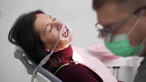 双眼鏡の男性歯科医は 現代の歯科クリニックで女性の歯を治療します 医者はベニヤを取付ける前に注射器が付いている歯の青いエッチングのゲルを加えます 歯科クラウンを設置するプロセス — ストック動画