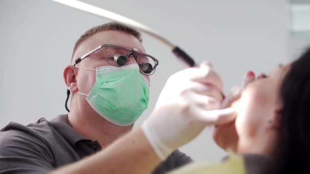 セラミックベニヤと王冠をインストールするための女性患者歯を準備する歯科双眼鏡の男性歯科医 マスクに集中した症候学者は 患者の檻を治療する 仕事中のドクター クローズアップ — ストック動画