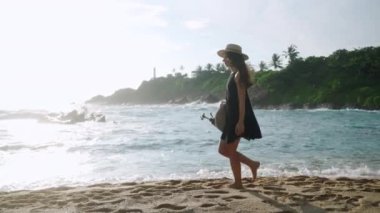 Sanatçı deniz kenarında yürür, dalgalar kıyıya vurur. Yaratıcı elbiseli, şapkalı, üç ayaklı, kumsalda gezen bir kadın. Kıyı bölgesinde içerik yaratan filmler. Güneşli tropikal manzara, seyahat blogcusu.