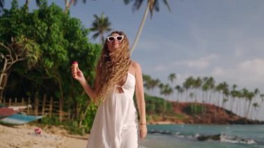 Sahil yürüyüşü, güneşli palmiye arkaplanı. Beyaz elbiseli bir kadın tropikal plajda dondurma tutarak geziniyor. Gülümsemeler dondurulmuş tatlıyı tadıyor, yaz neşesini somutlaştırıyor, tasasız tatilleri..