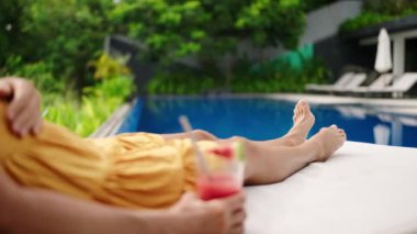Ayaklar havaya, ferahlatıcı içecekler, tropik yeşillikler arasında tatil yaşam tarzını somutlaştırıyor, özel villa ortamını. Rahat bir kadın havuz başında kokteyl ve sıcak güneş banyosu ile vakit geçirmeyi sever..