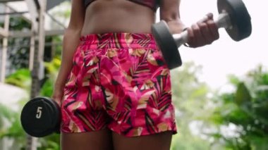 Canlı fitness kıyafeti kuvvet eğitimini arttırır. Formda bir kadın, tropikal bitkilerle çevrili bir spor salonunda halterlerle biseptik kıvrımlar yapar. Doğa içinde sağlıklı, aktif bir yaşam tarzı. Yavaş çekim.