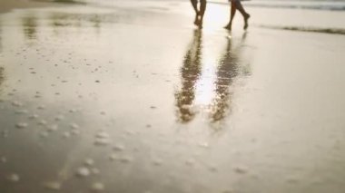 Gün doğumunda ıslak kumda iki kızın siluetinin yansıması. Yaz günbatımında gölgeler. Genç kadınlar tropik kıyı şeridi boyunca yürüyorlar. Uyumlu kızlar kumsalda yalınayak geziyor..