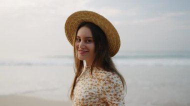 Çiçekli elbiseli, deniz kenarında gülümseyen, köpüklü su dalgaları olan çekici bir kız. Hasır şapkalı genç esmer kadın okyanus kıyısında yürüyor. Şık bir bayan turist flörtü, deniz kıyısında rahatlamak..