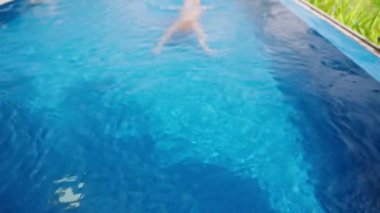 Lüks seyahat yaşam tarzı, güneşli villada dinlenme. Zarif bir kadın, tropikal bir havuzda mavi sudan çıkar. Boş zaman, keyif, bereketli bitkilerin olduğu yaz atmosferi. Yavaşla