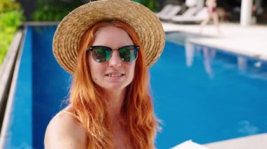 Rahat, güneşli tatil köyü atmosferi. Güneş gözlüklü kızıl saçlı kadın, saman havuz başında eğlenmeyi seviyor, kokteyllerini yudumluyor, arkadaşça video görüşmeleri yapıyor. Medya, teknoloji meraklısı akıllı telefondan milenyum anı. Yavaşla