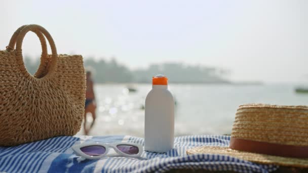夏季休闲活动的紫外线防护必需品 日常皮肤护理 海滩场景 有防晒霜瓶子和条纹毛巾 旁边有柳条袋 海太阳镜 背景是游泳者 — 图库视频影像