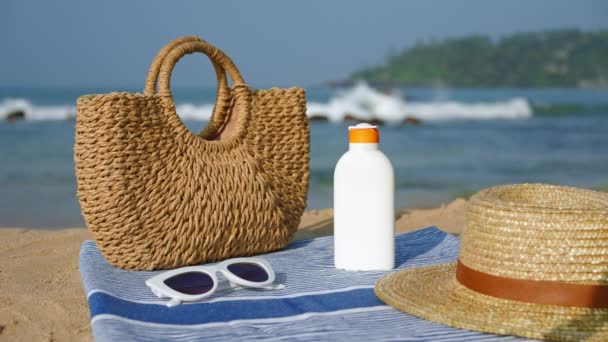 熱帯海岸の夏の皮の保護のための紫外線に対するプロダクト 日焼け止めの砂浜のボトルに配置されたビーチの必需品 スタイリッシュなわら帽子 タオルの上に日当たり ウィッカートート脇に — ストック動画
