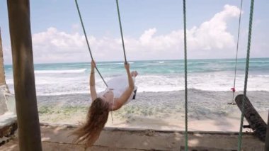 Turkuaz dalgalara bakarken rüzgar saçı kaldırıyor. Beyaz elbiseli kıvırcık saçlı bayan tropikal okyanusta salıncaktan hoşlanır. Sahilde egzotik bir an, özgürlük, güneşli bir günde mutluluk. Slomo