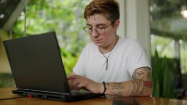 İkili olmayan girişimci odaklanıyor, dijital görevleri uzaktan yönetiyor, çeşitliliği somutlaştırıyor, dahil ediyor, çalışmada özgürlük sağlıyor. Transseksüel birey parlak bir kafede dizüstü bilgisayarda çalışır, yeşillik camdan görülür..