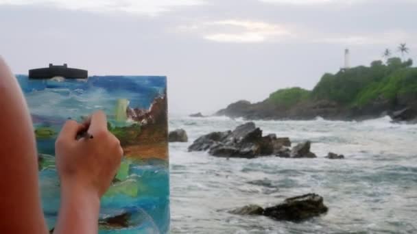 手握画笔 她融合了色彩 诠释了汹涌的海景 明显地体现了户外创作过程 女画家在岩石海岸边画画布 在暮色中捕捉波涛汹涌的波涛和灯塔的背景 — 图库视频影像