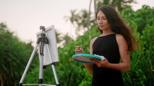 黒いドレスの女性は 海のビーチでイーゼルによって動作パレット ブラシを保持しています クリエイティブなアーティストが屋外のキャンバスに絵を描きます 画家は自然 熱帯の風景の中で芸術を創造する ホビーとレジャー活動 スローモ — ストック動画