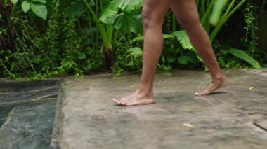 Vücudunun alt kısmı suya giriyor, ferahlatıcı yüzüşün tadını çıkarıyor. Çift ırklı kadın yeşil tropikal bitkilerle çevrili temiz havuza giriyor. Doğal ortam, boş zaman aktivitesi, sağlık inzivası konsepti. Yavaşla