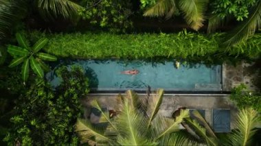 Huzurlu bir ortam, refah inzivaları, tek başına seyahat kaçışları, lüks tatil köyleri terfileri için mükemmel. Koyu tenli bir kadının yeşil havuzda rahatça yüzüşünü gösteren drone görüntüsü..