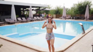 Güzel bir kadın kalıyor, poz veriyor, plaj restoranında yüzme havuzu ve güneşli yaz günlerinde arkadaşlarıyla flört ediyor. Mayolu ve şortlu bir model..