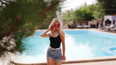 Sarı saçlı kadın kırmızı güneş gözlüğü, şort ve yüzme havuzunda güneş pansiyonları ve çam ağaçlarıyla fotoğraf çekmek için dans ediyor. Tatillerde poz vermek ve flört etmek.